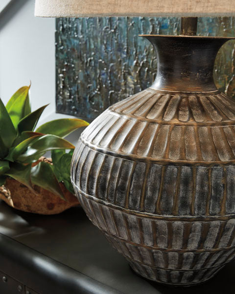 A close-up of the Ashley Magan Lamp L207354, featuring a large, ribbed ceramic base with a bronze-tone finish, sits on a table beside a green potted plant and a decorative bowl, with abstract artwork visible in the background.