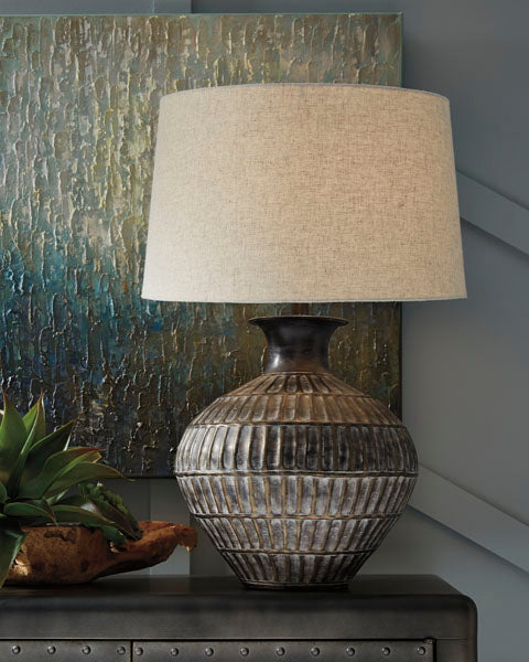 The Ashley Magan Lamp L207354, featuring a textured ceramic base with a bronze-tone finish and topped with a beige shade, sits on a dark surface next to a green plant, while an abstract multicolored painting hangs in the background.