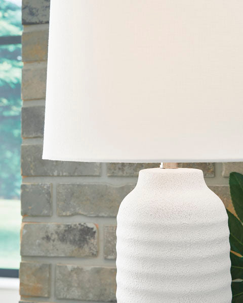 A close-up of the Ashley Vauville White Lamp L243674, featuring a sculptural white base and white shade, positioned in front of a gray brick wall with a window showcasing lush greenery outside.