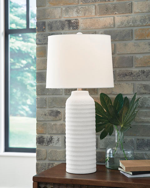 The Ashley Vauville White Lamp (L243674), featuring a textured ceramic sculptural base and a white shade, sits on a wooden table beside green leaves arranged in a glass vase. A brick wall and a window with views of greenery provide the backdrop.