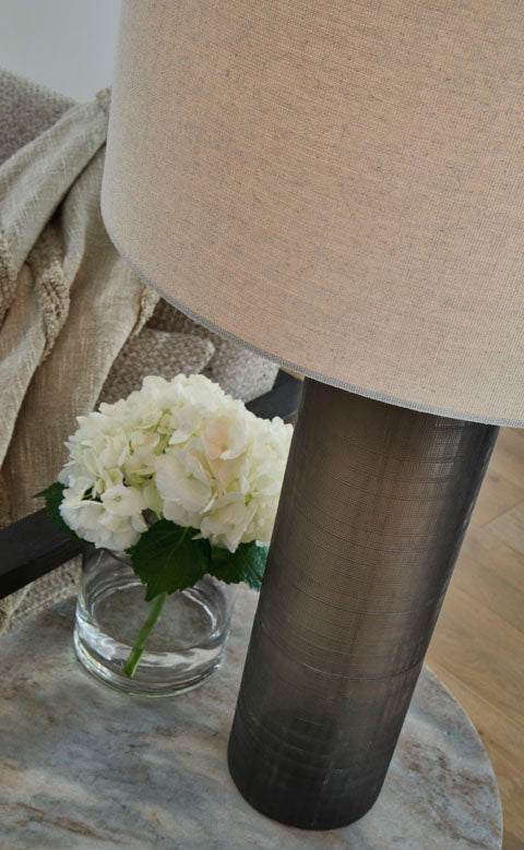 The Ashley Dingerly Lamp L430824, a cylindrical ombre table lamp with a neutral-colored drum shade and a frosted glass base, sits on a marble table next to a small glass vase of white hydrangea flowers.