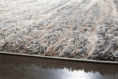Close-up of the Gizela Rug R404861 by Ashley, showcasing its soft, plush pile in elegant beige and gray tones. The rug features a thick, textured surface with upright fibers and is displayed on a dark wooden floor for a luxurious look.