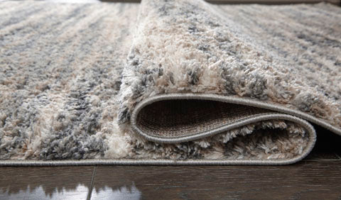 A close-up of the Ashley Gizela Rug R404861, featuring a beige and gray subtle striped pattern, is shown folded over on a dark wooden floor to highlight its plush texture and impressive thickness.