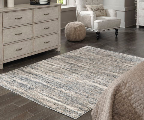 A cozy bedroom features a gray and beige striped design area rug with a plush pile on dark wood floors, with a dresser, armchair, knitted pouf, and part of a quilted bed visible. Light streams in through a window.