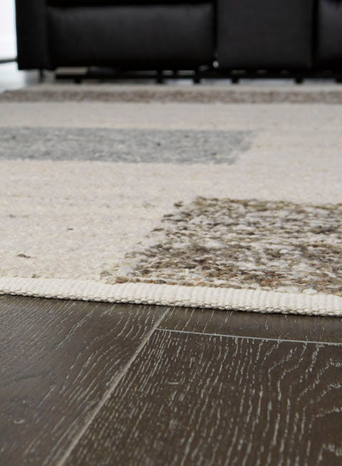 Ashley Barus Rug R406402, a handwoven area rug with a neutral-toned, abstract block pattern on a dark wood floor.