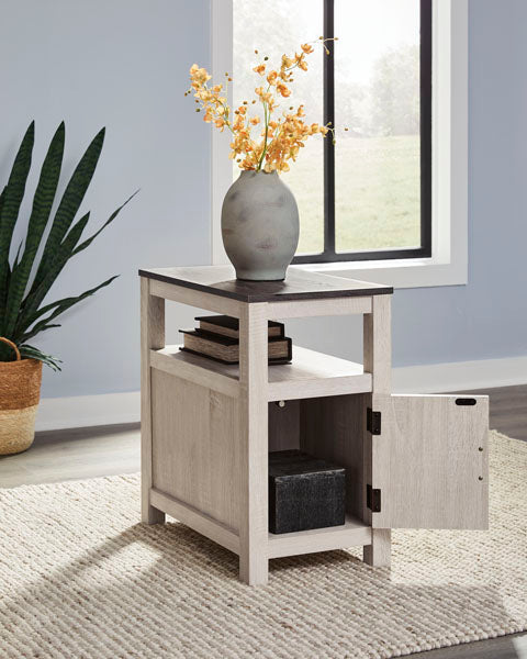 Fendenmore White Chairside End Table T190-217 featuring a light-colored wood frame with an open shelf, a lower storage cabinet with an open door, and a dark top holding a vase of yellow flowers.
