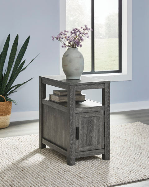 Fendenmore Gray Chairside End Table T190-317 in weathered gray featuring an open shelf with two books and a lower storage cabinet with a black handle, topped with a vase of purple flowers.