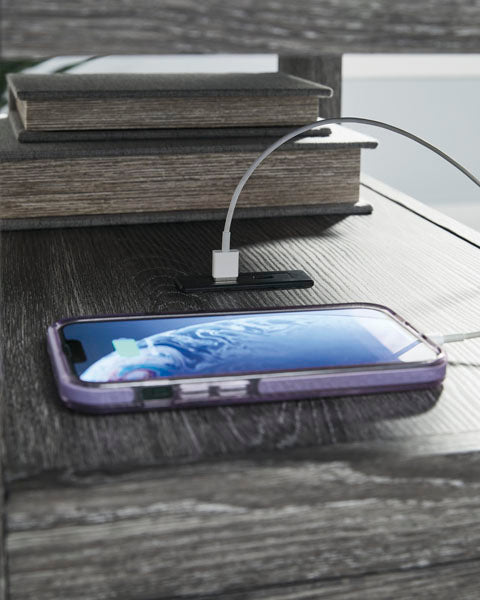 A Fendenmore Gray Chairside End Table T190-317 with a weathered gray finish, featuring a charging smartphone connected to a USB port on its surface and two books stacked in the background.