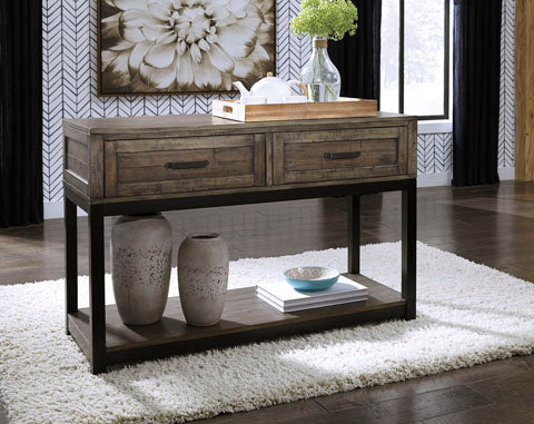 The Ashley Johurst Sofa Table T444-4 features a rustic wooden design with sturdy metal legs, sitting gracefully atop a white rug to add modern farmhouse charm to the space. On its surface rest a tray, a glass vase filled with flowers, and white teapots, while two decorative vases and a bowl are displayed on the lower shelf.