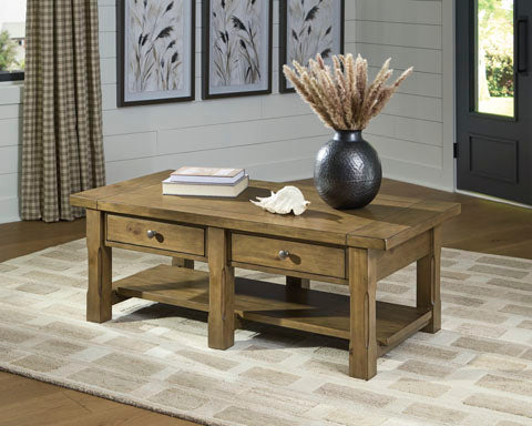 The Ashley Vandenmore Coffee Table T539-1, featuring two storage drawers and a farmhouse wooden design, sits atop a beige patterned rug. On the table are a book, a decorative shell, and a large vase filled with dried grasses. Framed art and plaid curtains provide a charming backdrop.