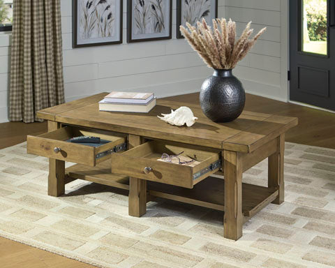 The Ashley Vandenmore Coffee Table T539-1 features a farmhouse-inspired wooden design with two open drawers for convenient storage of books and glasses. Styled with a vase of dried plants and a seashell on top, the table is set on a beige patterned rug in a cozy living room.