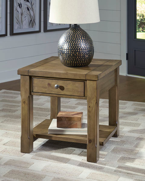 The Ashley Vandenmore End Table T539-3, featuring a farmhouse-inspired wooden design with a single drawer and lower shelf, rests atop a beige rug. Ideal for sofa-side storage, it supports a patterned decorative lamp and showcases two wooden boxes on the tabletop and shelf. Framed artwork and a nearby door provide an inviting backdrop.
