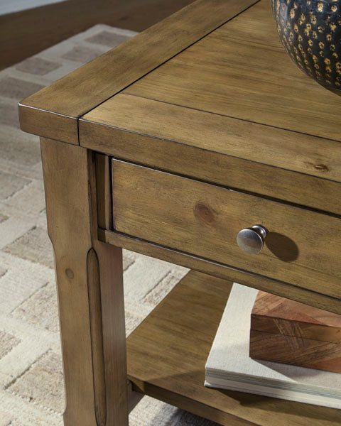 Close-up of the Ashley Vandenmore End Table T539-3, showcasing its farmhouse style with a drawer and metal knob for convenient sofa-side storage. The lower shelf neatly holds books, while part of a decorative bowl rests atop the table. A textured beige rug is partially visible underneath.