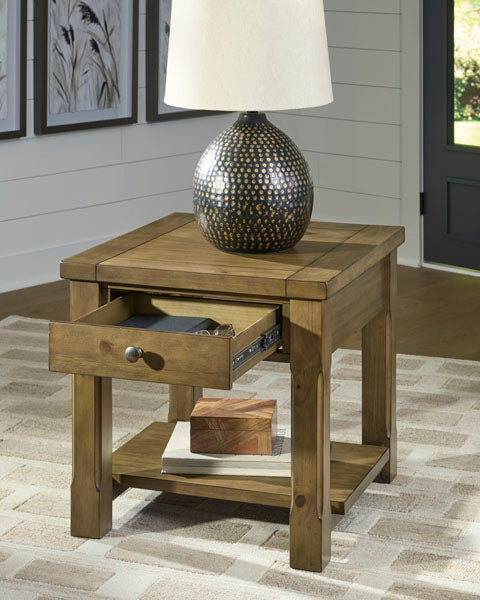 The Ashley Vandenmore End Table T539-3, featuring an open drawer and a lower shelf for books, sits on a patterned rug in a bright, modern room with a decorative lamp on top—providing convenient sofa-side storage.