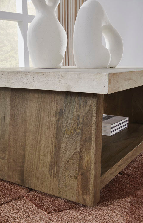The Brendalhouse Cocktail Table T642-1 by Ashley features a faux cement tabletop and wooden frame, with two abstract white sculptures and a stack of books placed on its lower shelf. A window with curtains can be seen in the background.