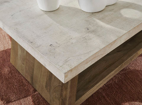 The Ashley Brendalhouse Cocktail Table T642-1 features a light-colored faux cement tabletop and a wooden base, positioned on a reddish patterned carpet. Three partially visible white round vases are arranged on the table.
