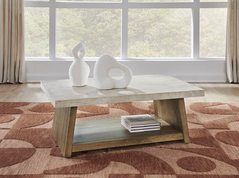 The Brendalhouse Cocktail Table T642-1 by Ashley features a rectangular, modern design with a light faux cement tabletop, wooden angled legs, and a convenient lower shelf for storing books. Two contemporary white sculptures are displayed on the table, while a large window and patterned rug provide an elegant backdrop.