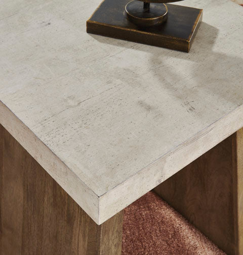A close-up of the Ashley Brendalhouse End Table T642-2 highlights its contemporary design, featuring a white faux cement tabletop and wooden legs. The table rests on a brown carpet and is topped with a dark, rectangular decorative object.