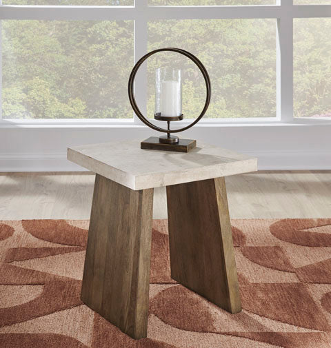 The Brendalhouse End Table T642-2 by Ashley features a square design with angled wooden legs and a faux cement tabletop. It is styled with a circular metal candle holder holding a white pillar candle, and is placed on a patterned rug near large windows overlooking lush greenery, adding contemporary flair to the space.