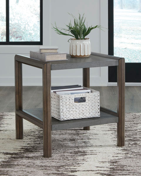 The Ashley Kallenny End Table T669-3, featuring a subtle mixed-material design with a gray wooden top, displays a potted plant and two books. Its lower shelf includes a white basket filled with magazines. Positioned on a patterned rug beside large windows, this small end table adds both function and style to the space.