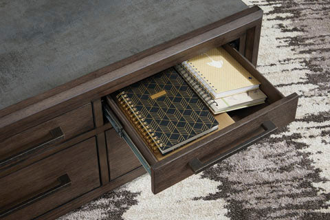 Several spiral notebooks with patterned covers are stacked neatly inside an open wooden drawer of the Ashley Kallenny Cocktail Table (T669-8), which rests on a textured rug and adds an industrial edge to the space.