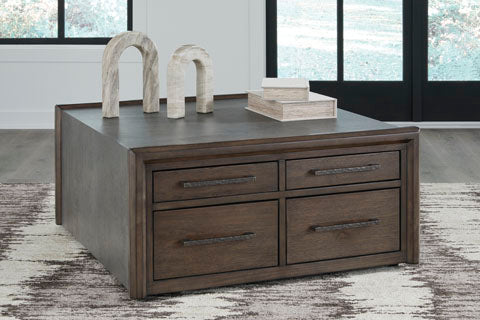 The Ashley Kallenny Cocktail Table (T669-8), featuring a square wooden design with four drawers, decorative arches, and stacked books on top, is set on a patterned rug in a modern living room with large windows. It brings an industrial edge to the space while serving as both a stylish cocktail table and functional centerpiece.