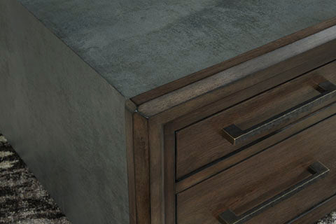 Close-up of the corner of the Ashley Kallenny Cocktail Table (T669-8), featuring a gray textured surface and metal handles atop a patterned rug, highlighting its industrial edge inspired by contemporary coffee table design.