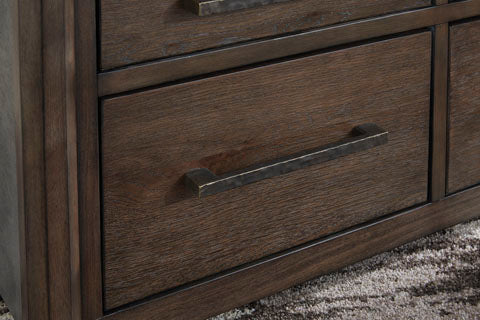 Close-up of the Ashley Kallenny Cocktail Table T669-8, featuring a dark wood finish and a long, rectangular metal handle that adds an industrial edge to the design. The table rests above a textured brown and gray carpet.