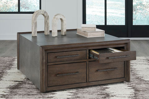 The Ashley Kallenny Cocktail Table (T669-8), a square wooden piece featuring four drawers—one partially open to reveal books inside—adds an industrial edge to this modern room. Decorative objects and stacked boxes rest atop the table, which sits on a patterned rug beneath expansive windows.