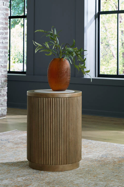 The Camdill End Table T808-6 by Ashley, featuring a ribbed cylindrical wooden design, holds an amber-colored vase with green leafy branches. The table is placed on a light rug in a contemporary living room near large windows and a brick wall, with natural light streaming in.