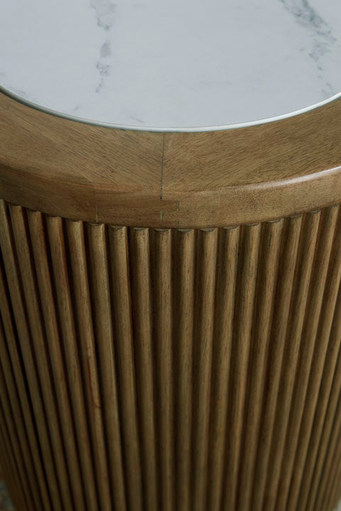 A close-up of the Ashley Camdill End Table T808-6, featuring a round wooden base with vertical grooves and a smooth faux Carrara marble top—an elegant accent perfect for a contemporary living room.