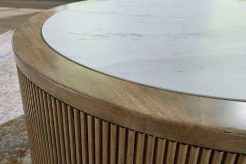 The Ashley Camdill Coffee Table (T808-8) features a round, contemporary design with a faux Carrara marble top and a light wood base accented by vertical slats; part of a rug can be seen nearby on the floor.