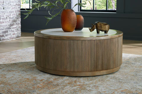 The Ashley Camdill Coffee Table T808-8 features a round wooden design with ribbed sides and a white top reminiscent of faux Carrara marble. Styled with two orange vases filled with greenery and a small wooden rhinoceros, it sits atop a patterned rug in a contemporary living room.