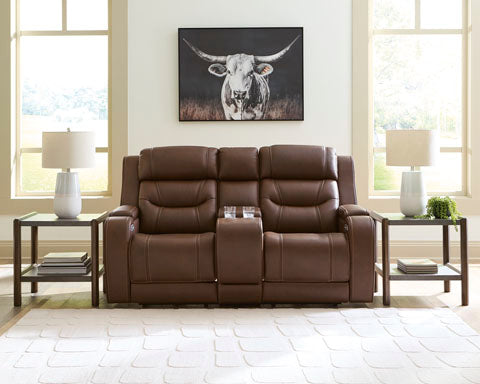 The Knightington Brown Power Reclining Loveseat (Ashley U1160218A) sits at the center of this inviting space, featuring a convenient center console. It’s flanked by end tables topped with white lamps, while a framed longhorn picture hangs above between two sunlit windows. The light rug underfoot and the cozy ambiance make it perfect for relaxing or movie nights—especially when complemented by a premium audio system.