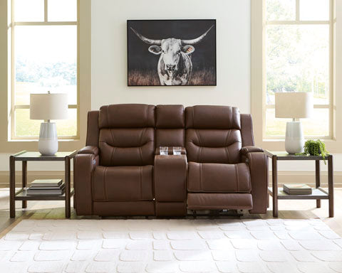 The Ashley Knightington Brown Power Reclining Loveseat (U1160218A) with a center console is positioned between two end tables topped with lamps, set in front of large windows. A framed cow picture is displayed on the wall above.