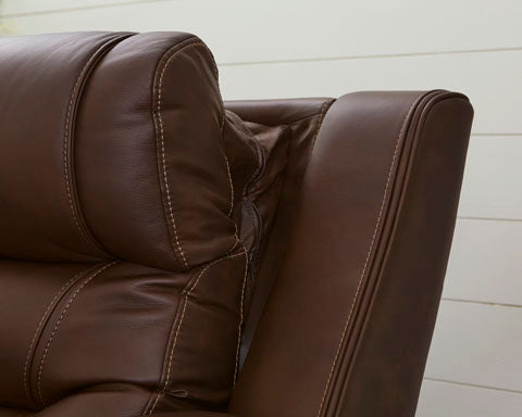Close-up of the Ashley Knightington Brown Power Reclining Loveseat (model U1160218A), featuring visible stitching and a detailed view of the headrest and armrest against a white wooden wall background—an ideal choice to elevate your home theater sofa arrangement.