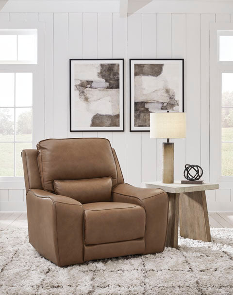 The DeepWakes Caramel Recliner U6260613 by Ashley, upholstered in top-grain leather with a rich caramel hue, rests on a shaggy rug beside a wooden side table adorned with a lamp and decor, all set in a bright room with white walls and abstract art displayed above.