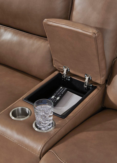 The DeepWakes Caramel Power Reclining Loveseat U6260618 by Ashley features rich brown top-grain leather upholstery, a central armrest equipped with two cup holders—one holding a glass of water—and an open storage compartment containing a remote control.