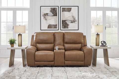 DeepWakes Caramel Power Reclining Loveseat Version: The Ashley DeepWakes Caramel Power Reclining Loveseat (U6260618) features rich caramel leather and a center console, flanked by two wooden side tables with lamps. A plant and decor enhance the space, while two abstract art pieces hang on the white shiplap wall behind the loveseat.