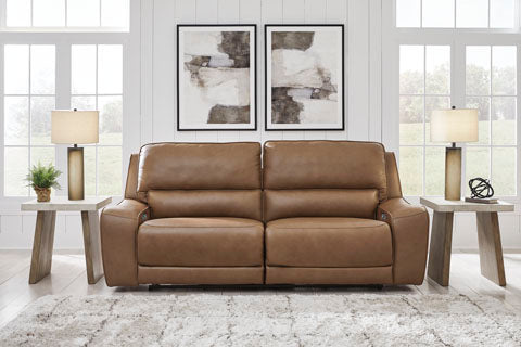 An Ashley DeepWakes Caramel Power Reclining Sofa (model U6260647) sits between two wooden end tables adorned with lamps and decor, arranged atop a soft patterned rug. Two abstract paintings are hung above the sofa, while large windows in the background fill the space with natural light.
