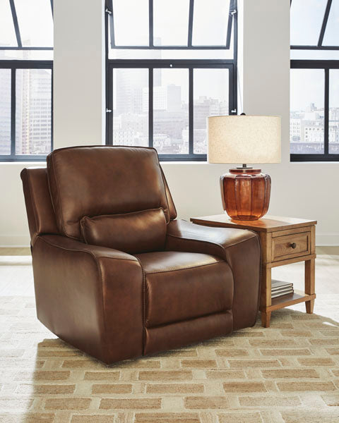 The Ashley DeepWakes Canyon Power Recliner U6260713 in brown top grain leather, featuring an adjustable headrest, sits beside a wooden side table topped with a brown glass lamp, all arranged on a patterned rug in front of large windows showcasing a cityscape view.