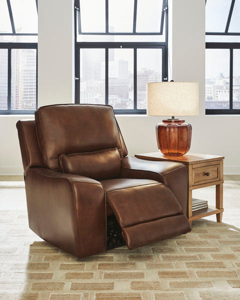 The DeepWakes Canyon Power Recliner U6260713 by Ashley, upholstered in brown top grain leather with an adjustable headrest and extended footrest, sits beside a wooden side table and amber lamp, positioned in front of large windows overlooking a cityscape.