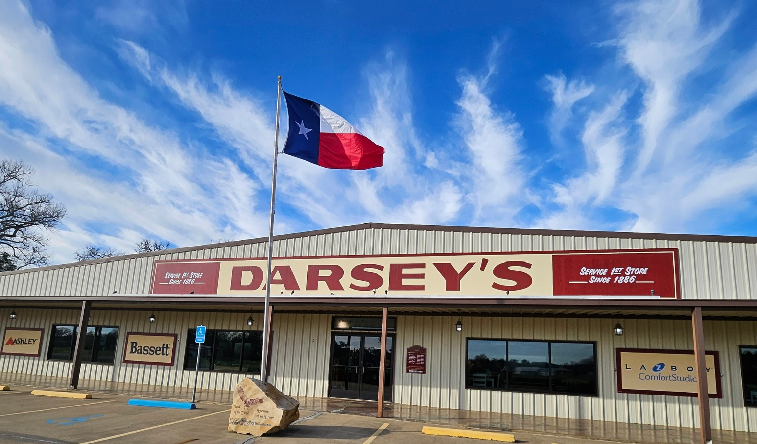 A Texas flag waves in front of Darseys furniture store, a large metal building with signs for brands like Bassett and La-Z-Boy under a blue sky with wispy clouds.