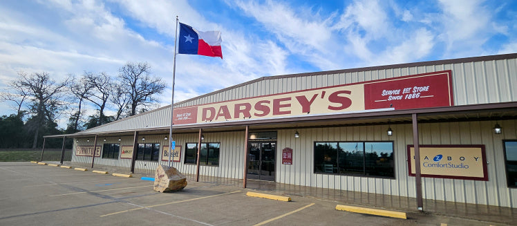 A large tan and brown building with a sign reading “Darsey’s” and a Texas flag flying in front. The storefront has several windows and signs, with empty parking spaces and a large rock nearby.